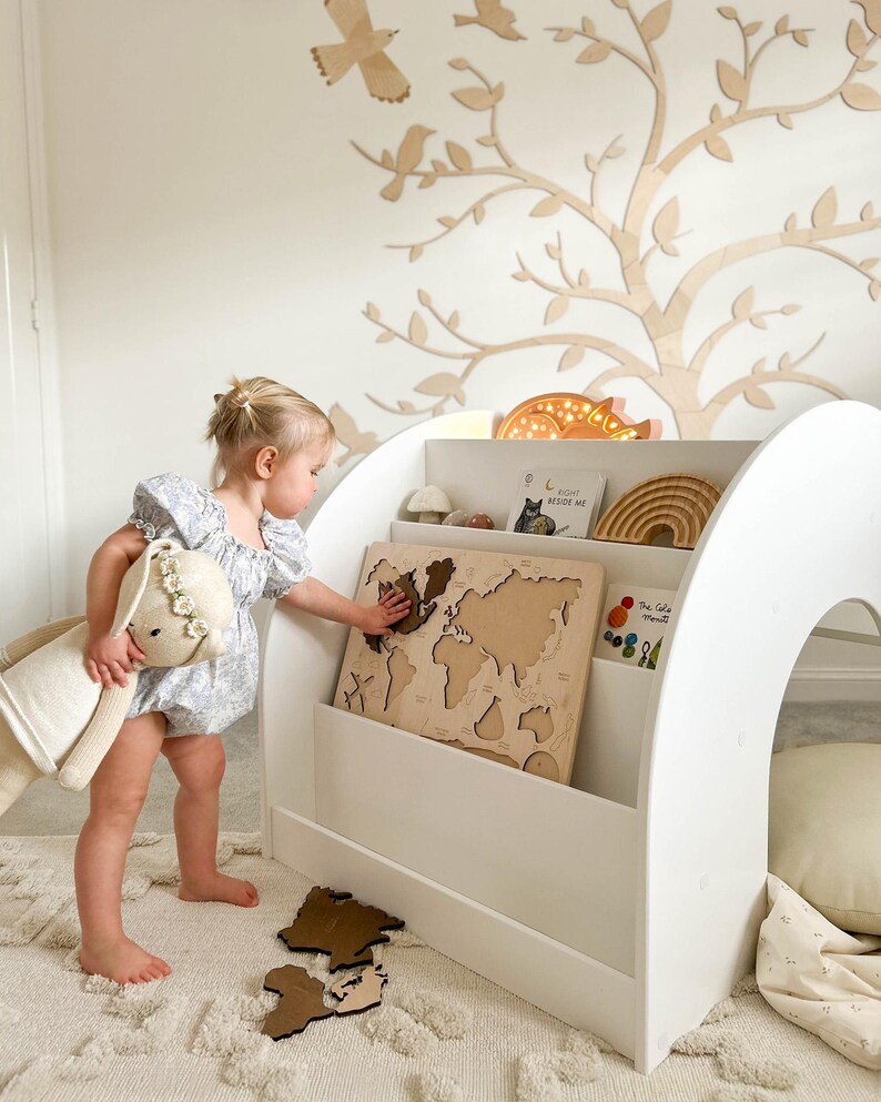 May include: A white wooden bookshelf with a built-in playhouse, featuring a world map puzzle and a child playing with the puzzle pieces. The bookshelf has three shelves and a curved top. The playhouse has a small opening and a soft cushion inside.