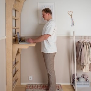 May include: A wooden climbing wall with a desk attachment. A person stands at the desk, using a laptop. The wall is mounted on a light-colored wall. A rug with a geometric pattern is on the floor.
