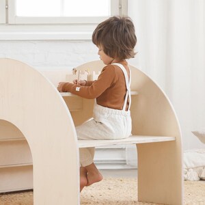 May include: A child sits at a wooden play table and chair set. The table and chair are made of light wood and have a rounded, arched design. The child is wearing a brown long-sleeved shirt and white overalls. The child is playing with wooden blocks.