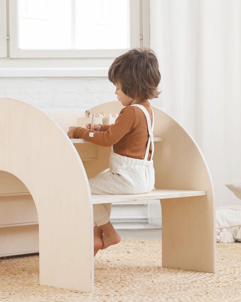 May include: A child sits at a wooden play table and chair set. The table and chair are made of light wood and have a rounded, arched design. The child is wearing a brown long-sleeved shirt and white overalls. The child is playing with wooden blocks.
