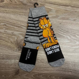 Könnte beinhalten: Graue Socken mit schwarzen Querstreifen und der Cartoon-Figur Garfield. Auf den Socken steht der Text "DON'T KNOW DON'T CARE" auf der Zehenpartie. Die Socken sind aus weichem Material.