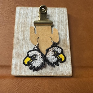 May include: Embroidered eagle head earrings with white, yellow, and black details. The earrings are displayed on a wooden tag with a brass clip. The eagle heads have a detailed, textured appearance.