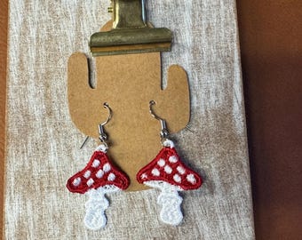 Op de afbeelding: Een paar paddenstoelvormige oorbellen met rode kappen en witte stippen. De oorbellen hebben witte stelen en zilverkleurige haken. Ze worden gepresenteerd op een houten oppervlak met een metalen clip.