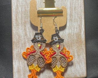 May include: A pair of embroidered turkey earrings with orange, yellow, brown, and black details. Each earring features a turkey wearing a pilgrim hat. The earrings are displayed on a wooden background with a gold clip.