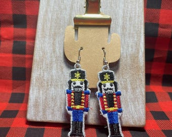 Puede incluir: Pendientes colgantes bordados con un diseño de soldado cascanueces. Los pendientes son negros, rojos, azules y amarillos. Se muestran en una tabla de madera con un clip dorado, sobre un fondo de cuadros rojos y negros.