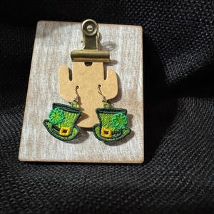 Op de afbeelding: Geborduurde groene kabouterhoed oorbellen met gele accenten en een klavertje vier. De oorbellen worden gepresenteerd op een houten houder met een messing clip. De achtergrond is een donkere, getextureerde stof.