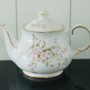 May include: A white teapot with a floral design in shades of pink, blue, and green. The teapot has a gold trim around the lid and spout.