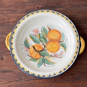Peut inclure: Un plat de service en céramique blanche avec deux poignées jaunes. Le plat est décoré d'un motif peint à la main de fruits d'oranger sur une branche avec des feuilles vertes et des fleurs roses. Le texte "RG Vietri" est imprimé au fond du plat.
