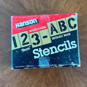 Peut inclure: Une boîte rouge, noire et dorée de pochoirs en laiton réutilisables Reese's Interlocking 123-ABC. La boîte est étiquetée "The C.H. Hanson Company" et "Manufactured by C.H. Hanson Company - Franklin Park, IL."