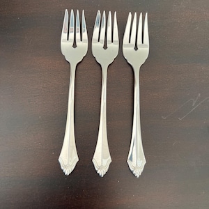 Peut inclure: Trois fourchettes argentées brillantes, chacune avec quatre dents, disposées verticalement sur une surface brun foncé. Les fourchettes présentent un motif décoratif à la base du manche. Les dents sont espacées uniformément.