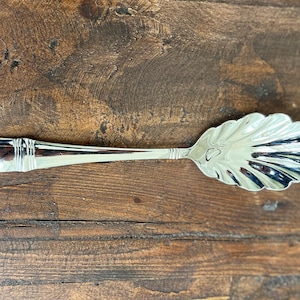 May include: A silver sugar spoon with a scalloped edge. The spoon is lying on a wooden surface.