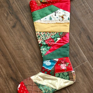 Peut inclure: Une chaussette de Noël en patchwork avec un motif rouge, vert, or et bleu. La chaussette est faite de différents tissus avec divers motifs de Noël, notamment des flocons de neige, des rennes et des sapins de Noël.