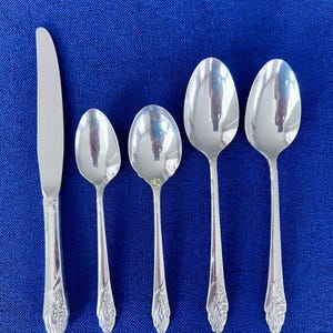 May include: A collection of silver-coloured cutlery, comprising a knife and four spoons, displayed on a blue surface. The knife and spoon handles have a decorative floral pattern. The spoons are of varying sizes.