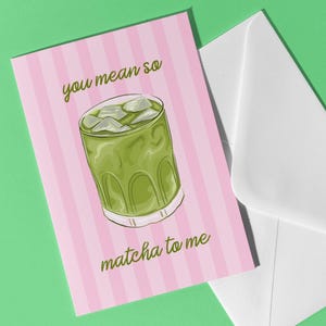 May include: A pink card with vertical white stripes and the text "you mean so matcha to me" next to a white envelope on a green background. The card features a drawing of a matcha drink with ice.