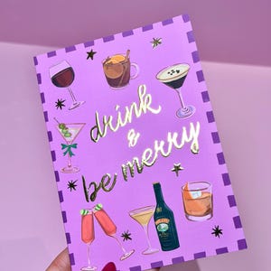 May include: A purple greeting card with gold foil lettering that reads "drink & be merry." The card features illustrations of various cocktails, including martinis, wine, and champagne flutes. Black star accents and a dotted border complete the design.