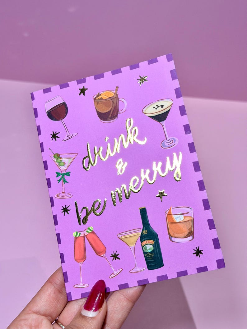 May include: A purple greeting card with gold foil lettering that reads "drink & be merry." The card features illustrations of various cocktails, including martinis, wine, and champagne flutes. Black star accents and a dotted border complete the design.