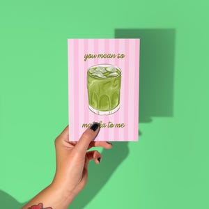 May include: A greeting card featuring a matcha drink illustration. The card has a pink and white striped background and the words "you mean so" and "matcha to me". The card is held against a green background.