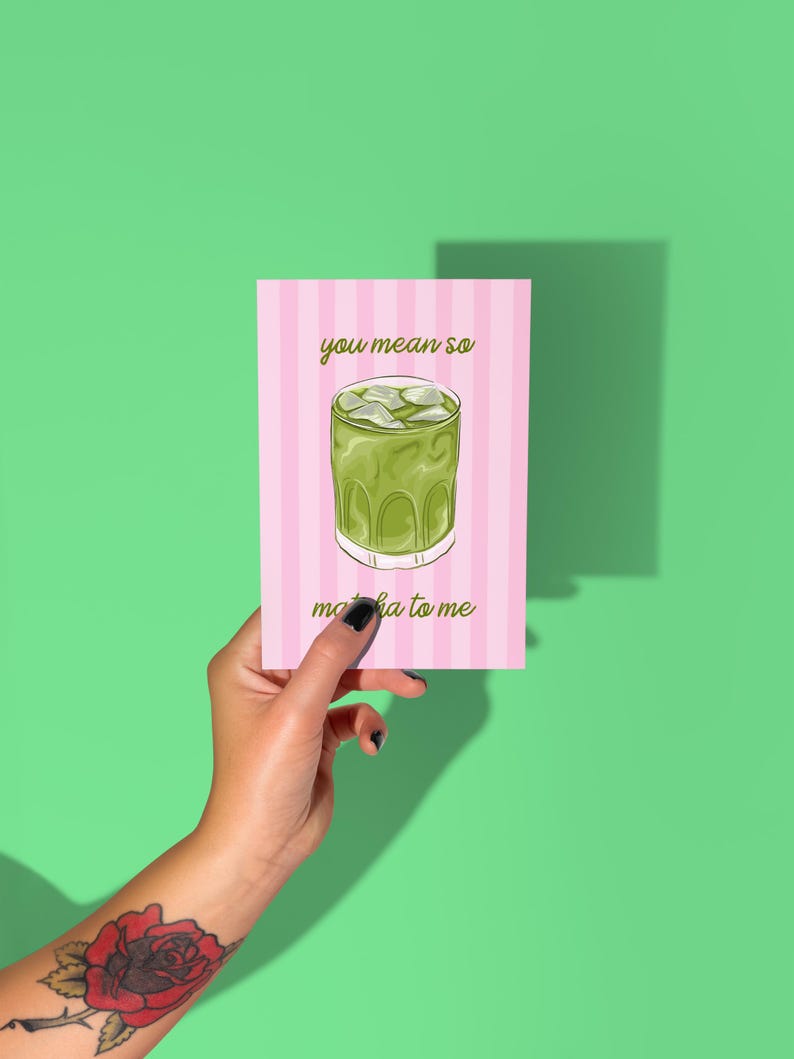 May include: A greeting card featuring a matcha drink illustration. The card has a pink and white striped background and the words "you mean so" and "matcha to me". The card is held against a green background.