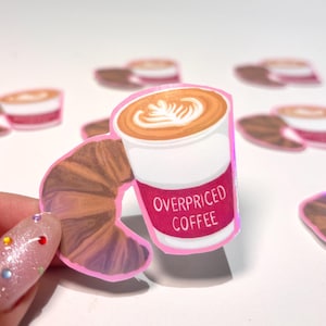Könnte beinhalten: Ein Aufkleber mit einer Kaffeetasse mit einem Croissant-Griff. Der Kaffee hat Latte-Art und die Tasse hat ein rosa Band mit den Worten "OVERPRICED COFFEE". Im Hintergrund befinden sich weitere Kaffeetassen-Aufkleber.