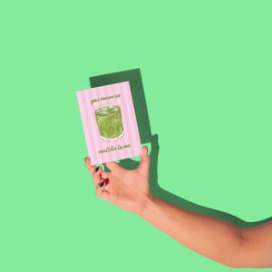 May include: A pink greeting card with the text "you mean so matcha to me" and an illustration of a matcha drink. The card has a pink and white striped background. The card is held against a green background.