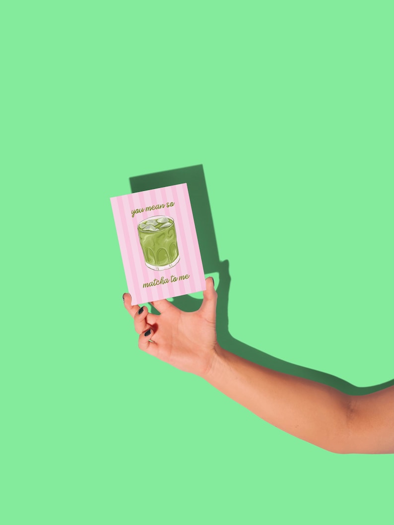 May include: A pink greeting card with the text "you mean so matcha to me" and an illustration of a matcha drink. The card has a pink and white striped background. The card is held against a green background.