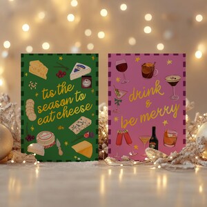 May include: Two festive greeting cards with holiday themes. One green card says "tis the season to eat cheese" with cheese illustrations. The other pink card says "drink & be merry" with drink illustrations. Decorative lights and ornaments surround the cards.
