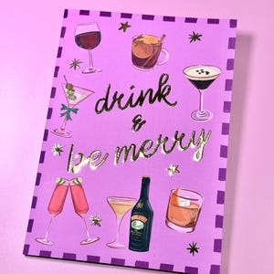 May include: A purple greeting card with gold lettering that reads "drink & be merry." The card features illustrations of various cocktails, including a martini, wine, and a bottle of Bailey's.