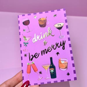 May include: A purple greeting card with illustrations of various cocktails and the words "drink & be merry" in gold lettering. The card features a border of purple squares and star accents.