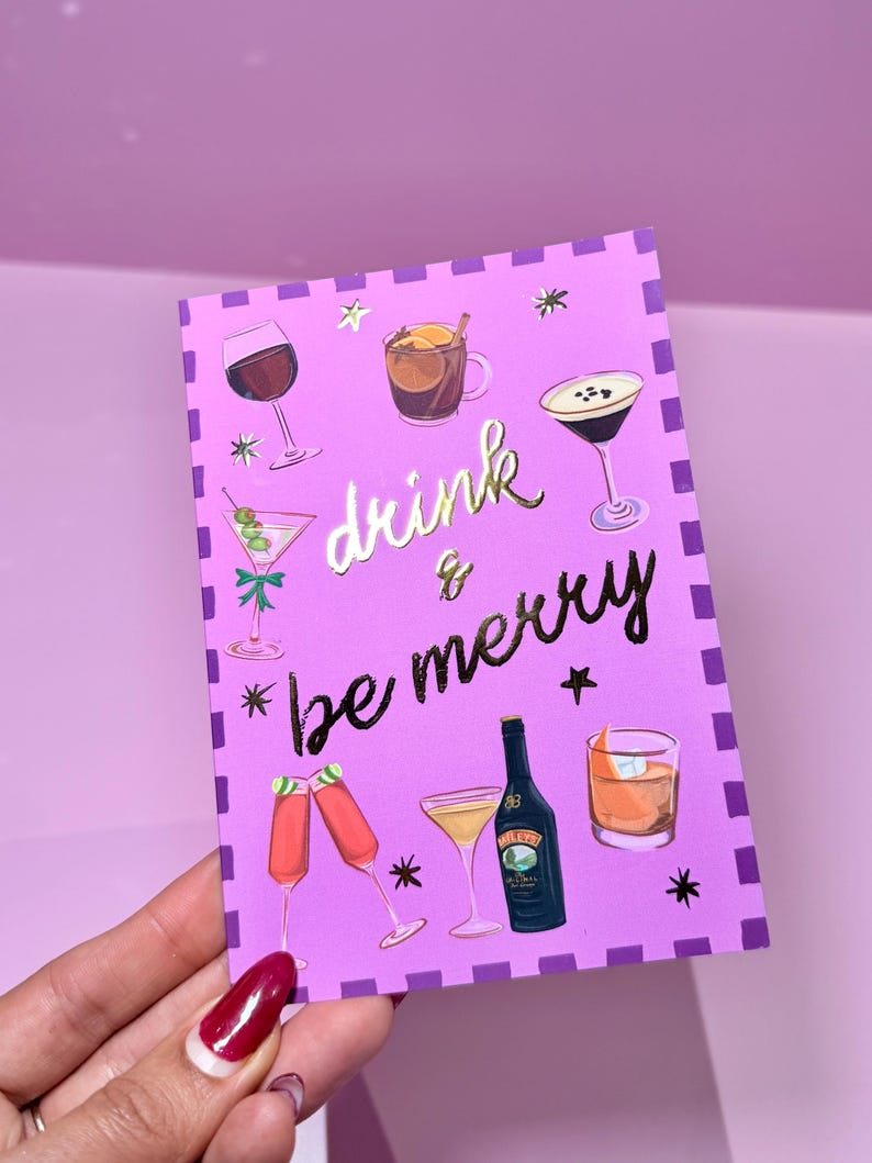 May include: A purple greeting card with illustrations of various cocktails and the words "drink & be merry" in gold lettering. The card features a border of purple squares and star accents.