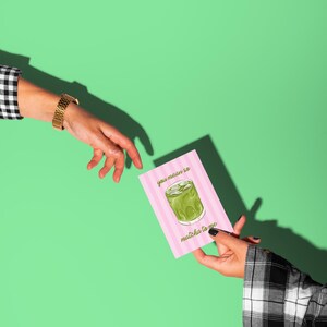 May include: A pink card with vertical stripes and the text "you mean so matcha to me" is held by a person. The card features a matcha drink illustration. A person's hand with a gold watch reaches toward the card against a green background.