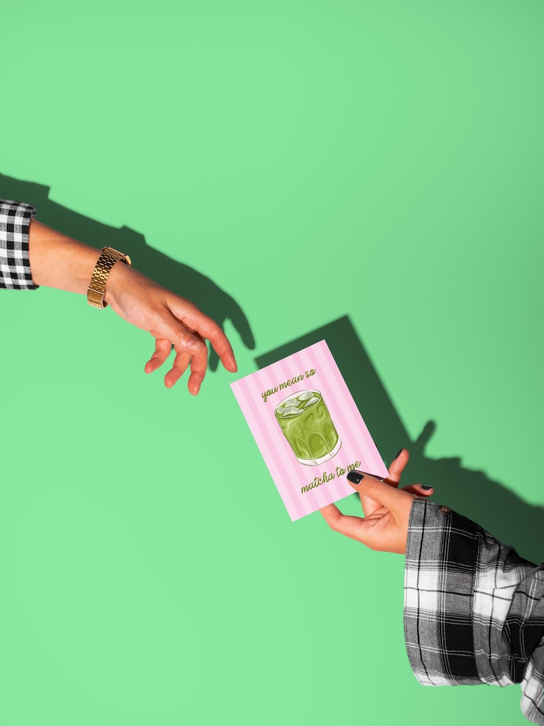 May include: A pink card with vertical stripes and the text "you mean so matcha to me" is held by a person. The card features a matcha drink illustration. A person's hand with a gold watch reaches toward the card against a green background.