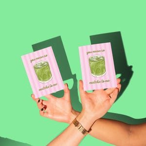 May include: Two pink cards with a matcha drink illustration and the text "you mean so matcha to me" are held up against a green background. The cards have a striped design. A gold bracelet is visible.
