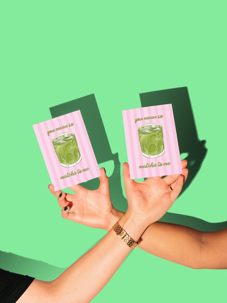 May include: Two pink cards with a matcha drink illustration and the text "you mean so matcha to me" are held up against a green background. The cards have a striped design. A gold bracelet is visible.