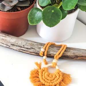 Peut inclure: Un collier en macramé jaune moutarde avec un anneau en bois et des franges. Le collier est posé sur une branche de bois sur un fond blanc.