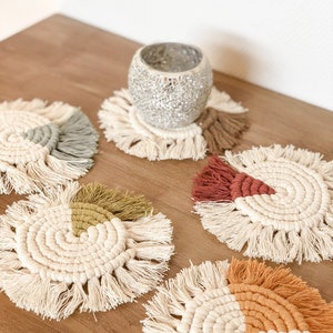 Peut inclure: Cinq dessous-de-verre ronds en crochet avec des franges, chacun avec une section de couleur différente. Les dessous-de-verre sont sur une surface en bois avec un bougeoir argenté au centre.