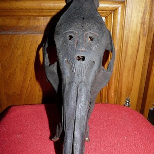 May include: A black animal skull with a carved face on the front. The skull is made of a dark material and has a rough texture. The face has two eyes, a nose, and a mouth.