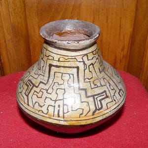 May include: A handcrafted, ceramic vase with a rounded body and a narrow neck. The vase features a cream-colored background with a repeating geometric pattern in black. The rim is a dark brown color. The vase is displayed on a red surface.