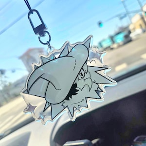 Può includere: Ciondolo per auto in acrilico trasparente con un personaggio dei cartoni animati. Il ciondolo ha un viso bianco, occhi neri e un bordo a forma di stella appuntita. È appeso a un cordino nero e a un fermaglio in metallo, per lo specchietto retrovisore.