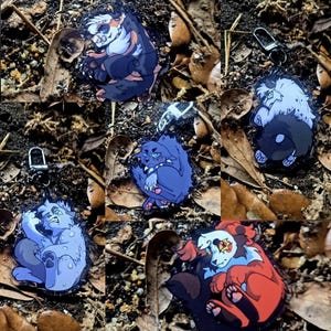 Könnte beinhalten: Eine Sammlung von fünf Acryl-Schlüsselanhängern mit stilisierten Illustrationen anthropomorpher Kreaturen in verschiedenen Farben wie Blau, Weiß, Schwarz und Rot, jeweils ausgestattet mit einem Metall-Karabinerverschluss.