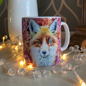 May include: A white ceramic mug featuring a colourful illustration of a fox's face surrounded by pink and red flowers. The mug has a white handle.
