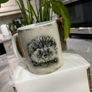 May include: A white ceramic mug with a detailed illustration of a hedgehog. The mug has a distressed, vintage-style finish and a white handle. The hedgehog is depicted in shades of grey and black, with a detailed, textured appearance.