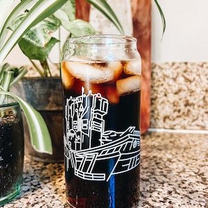 May include: A glass with a black and white graphic of a city skyline on it. The glass is filled with ice and a dark liquid, possibly coffee or iced tea.