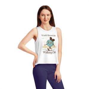 Puede incluir: Top blanco sin mangas de corte corto con un gráfico de una Tierra de dibujos animados que lleva una diadema de orejas de Mickey Mouse y camina por un camino a cuadros. El texto "World Showcase Walking Club" está impreso debajo del gráfico.