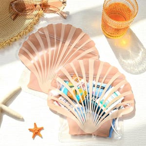 Op de afbeelding: Twee schelpvormige, doorzichtige plastic zakjes gevuld met snacks. De zakjes zijn lichtroze en hebben een ritssluiting. Een strohoed, zonnebril, een glas sinaasappelvloeistof, een zeester en een kleine oranje ster zijn ook zichtbaar.