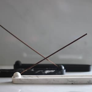 May include: A white marble incense holder with a round, white marble ball at one end. Two incense sticks are crossed over the holder. The holder is sitting on a black marble surface.