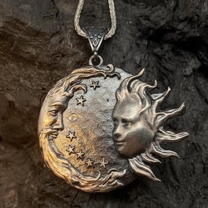 May include: A silver-toned circular pendant featuring a relief design of a crescent moon face on the left and a sun face on the right, separated by a field of small stars. The pendant hangs from a thin silver-toned chain link necklace.