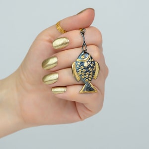 K&ouml;nnte beinhalten: Ein goldener Fisch-Anh&auml;nger mit schwarzen Details, der an einer goldenen Kette h&auml;ngt. Der Anh&auml;nger wird von einer Hand mit goldener Manik&uuml;re gehalten.
