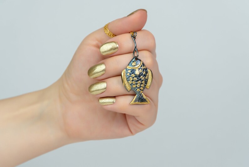 K&ouml;nnte beinhalten: Ein goldener Fisch-Anh&auml;nger mit schwarzen Details, der an einer goldenen Kette h&auml;ngt. Der Anh&auml;nger wird von einer Hand mit goldener Manik&uuml;re gehalten.