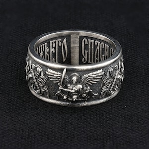 Puede incluir: Un anillo de plata con detalles intrincados. El anillo presenta un diseño central de una figura alada sosteniendo una espada, rodeada de patrones ornamentados y texto cirílico. El anillo tiene una banda ancha y un acabado pulido.