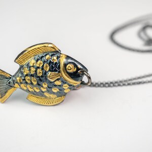 K&ouml;nnte beinhalten: Eine Halskette mit einem goldenen und schwarzen Fisch-Anh&auml;nger an einer schwarzen Kette. Der Fisch ist detailliert mit Schuppen und einem kleinen Auge.